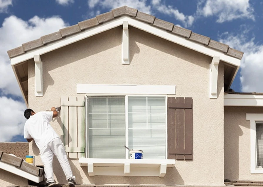 Homme en tenue de peintre avec casquette jaune appliquant de l'enduit ou peinture sur mur intérieur blanc près d'une fenêtre